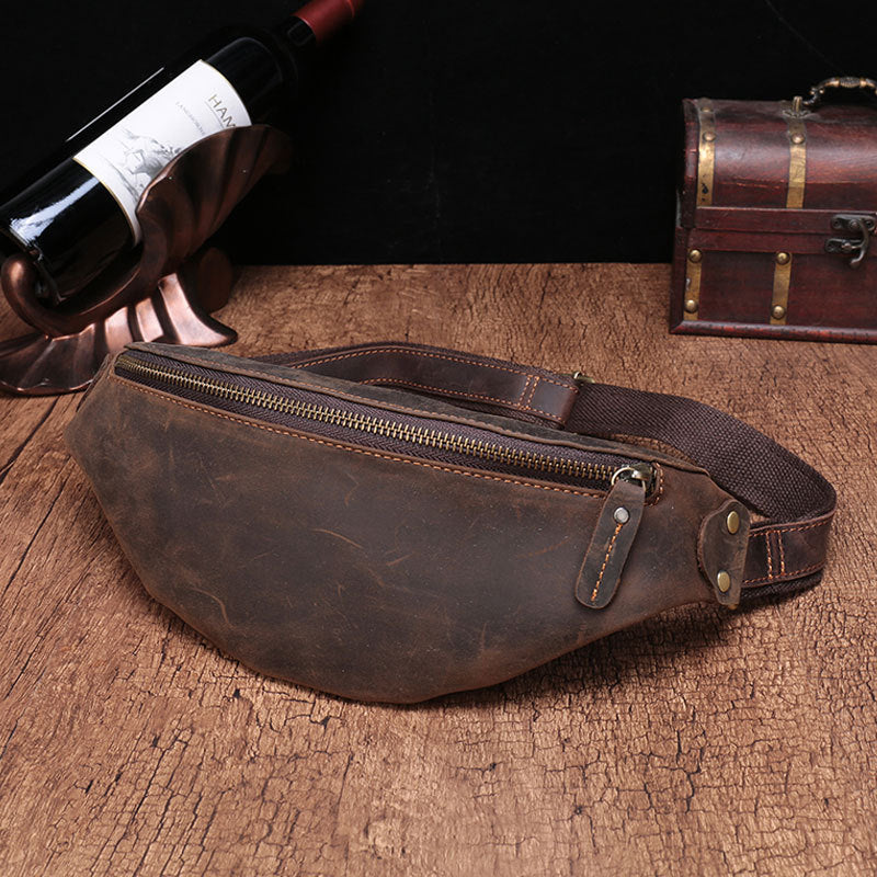 Rugged leather men's cell phone belt bag with zippered compartment, against a dark backdrop with a wine bottle and antique treasure chest.