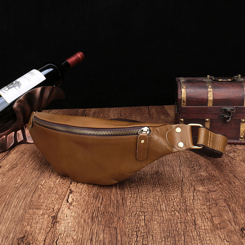 Camel-colored leather men's phone belt bag, with a zipper compartment, placed on a rustic wooden surface alongside a wine bottle and an old trunk.