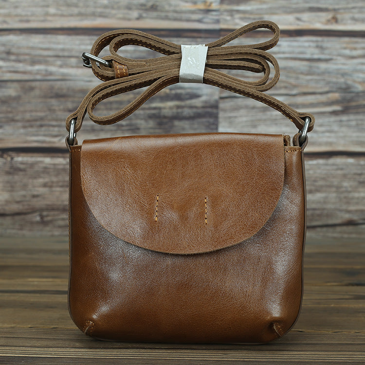 Tan leather crossbody bag with adjustable strap, simple design, and small front pocket detail, placed on rustic wooden background.