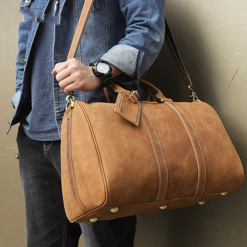 Vintage distressed leather travel luggage bag in camel color, carried by a man wearing a denim jacket and wristwatch.