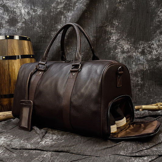 Large brown leather travel bag with handles and detachable strap, placed on a rustic wooden surface with a wooden barrel in the background.